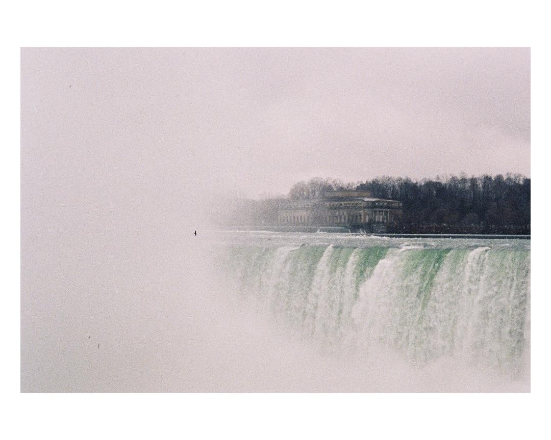 A wide shot of Niagara Falls shows a building and trees in the background, with mist obscuring the left side of the frame.