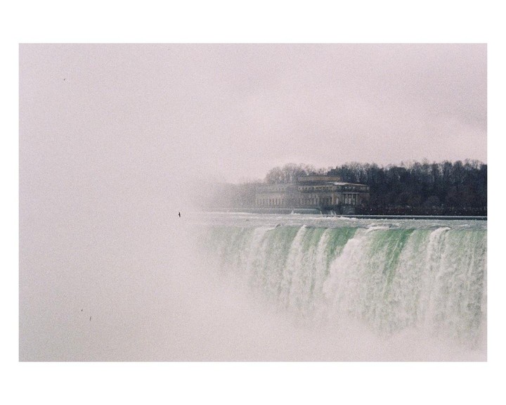 A wide shot of Niagara Falls shows a building and trees in the background, with mist obscuring the left side of the frame.