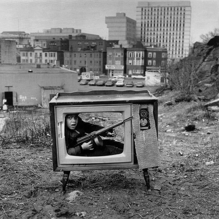 A boy with a toy gun is inside a television set in a city.