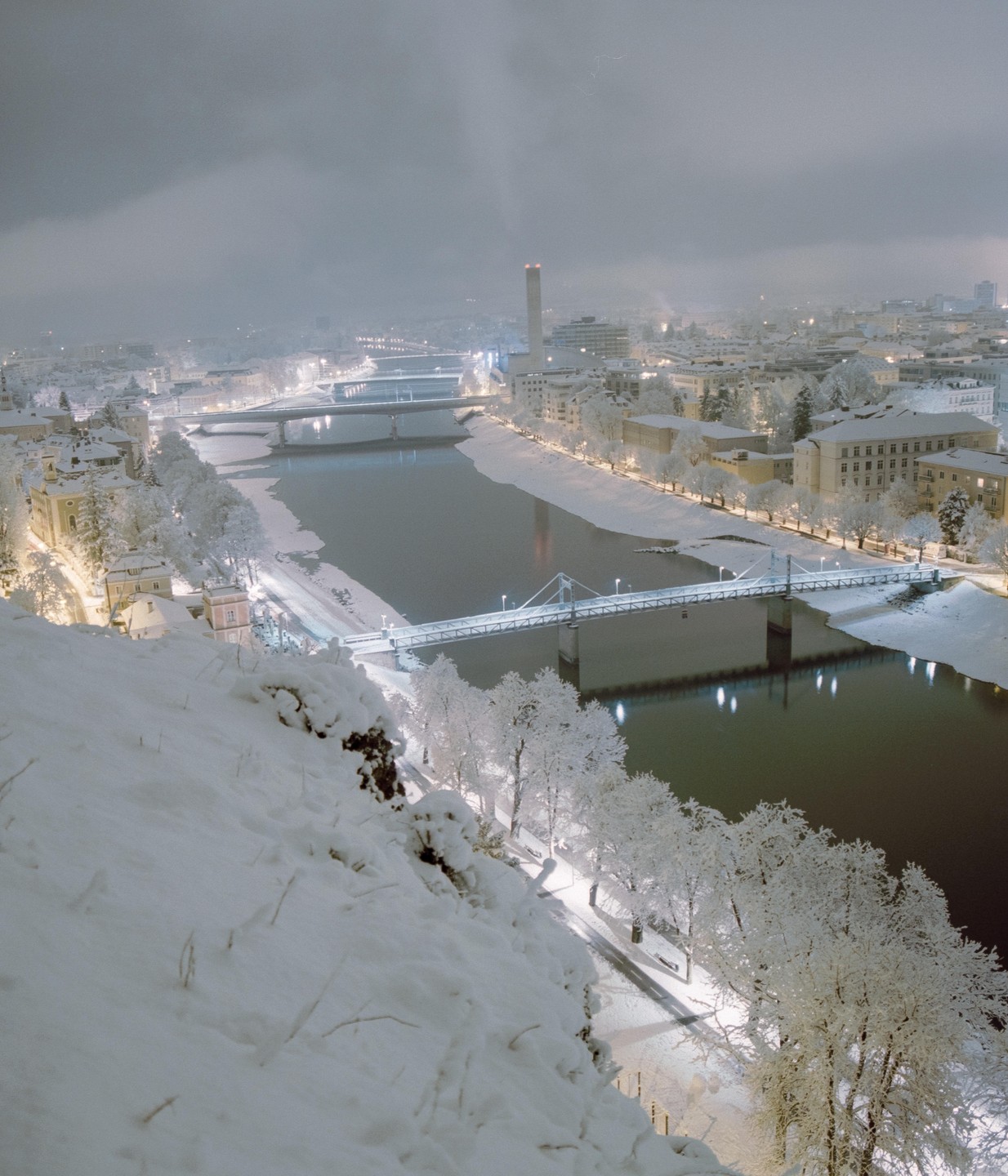 A snow-covered city with a river and bridges is illuminated at night.