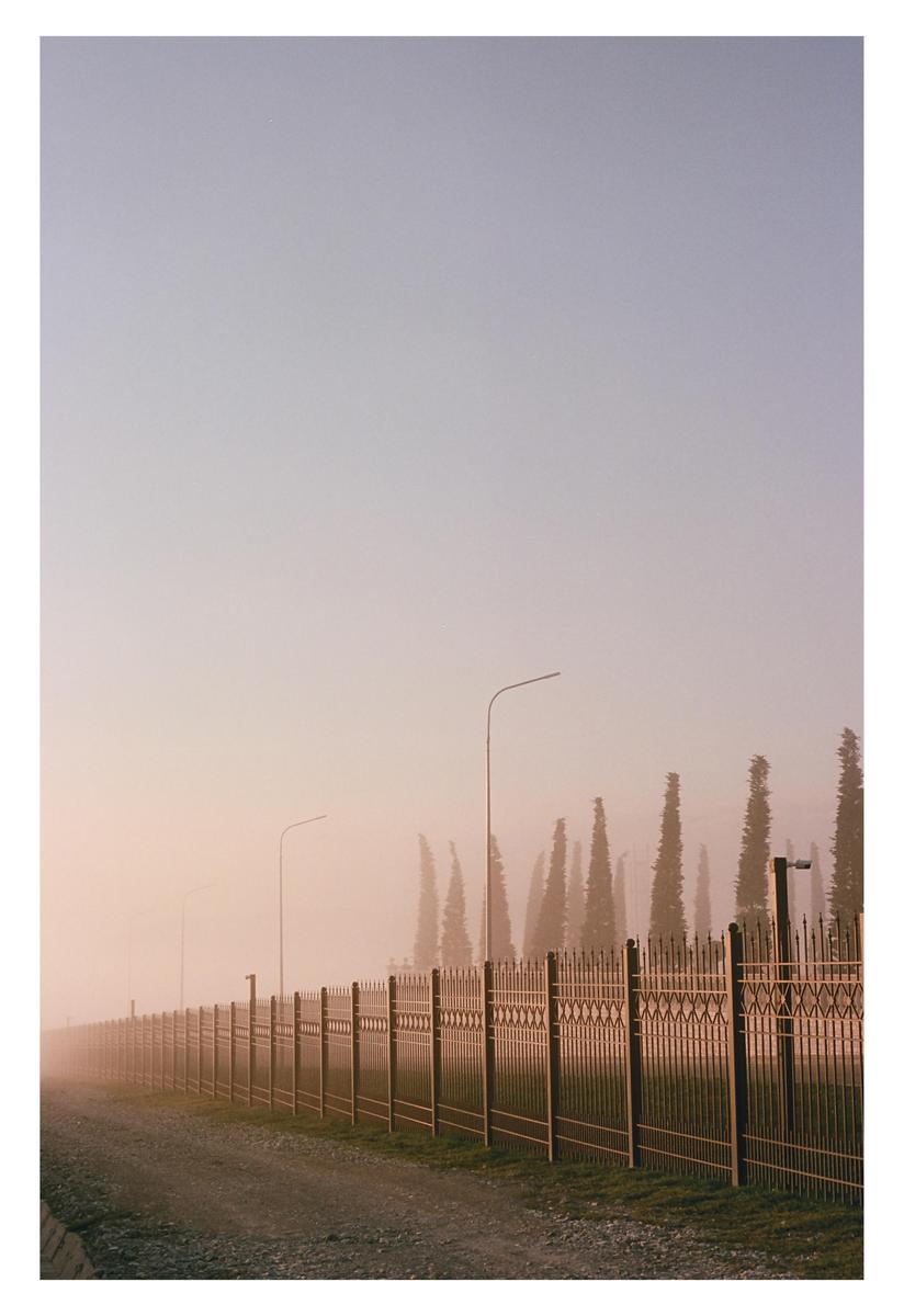 A long metal fence runs alongside a dirt road with tall, thin trees and streetlights in the background, all shrouded in fog.