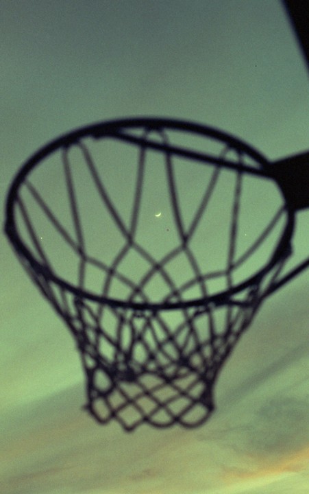 A crescent moon is visible through the net of a basketball hoop against a green sky.