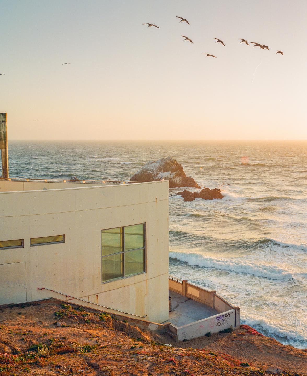 A flock of birds flies over a building on a cliff overlooking the ocean at sunset.