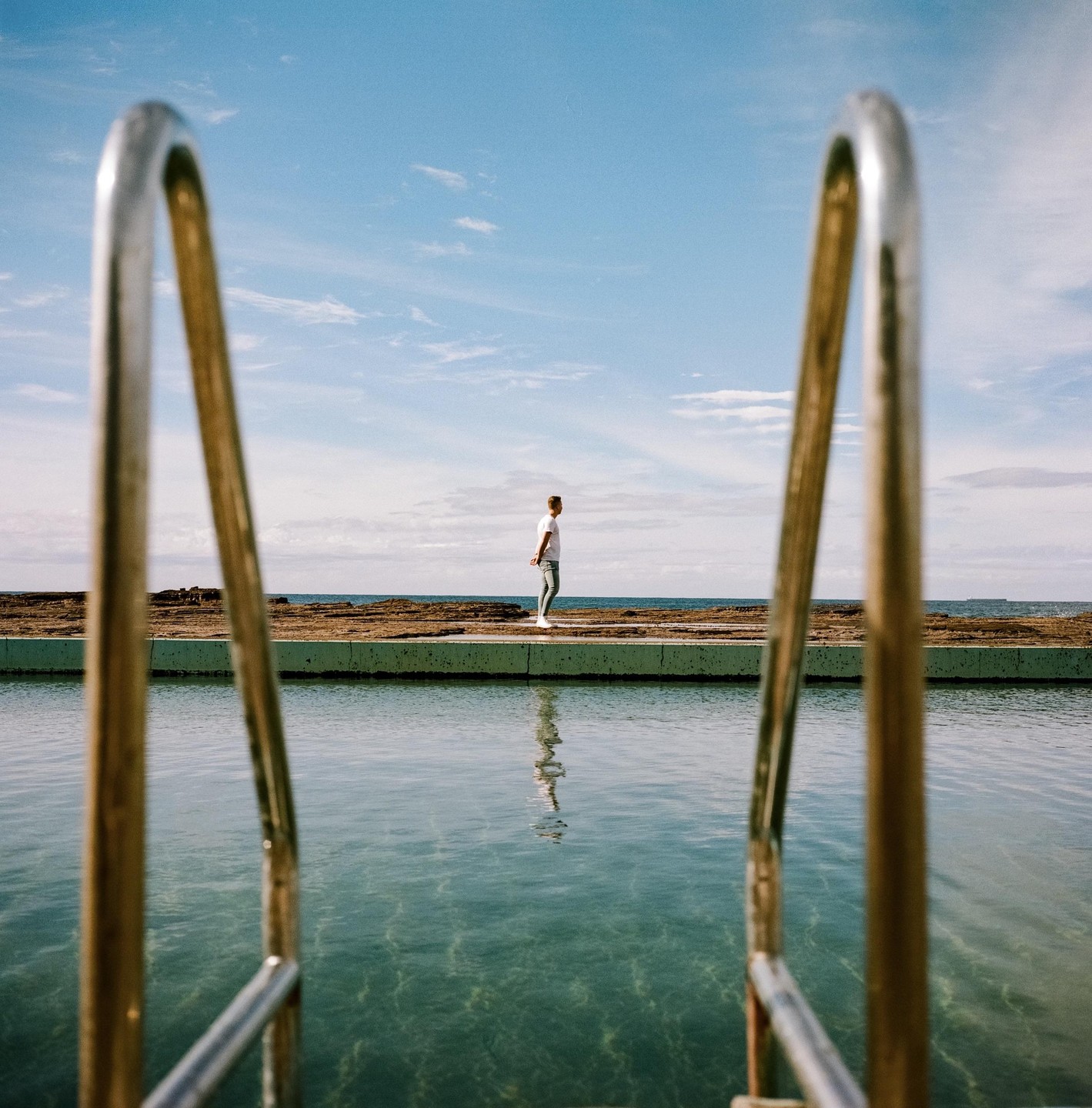 A man stands at the edge of an ocean pool, looking out at the sea, framed by the pool's metal ladder.