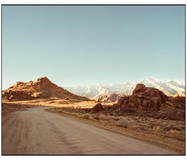 A dirt road winds through a rocky, arid landscape with snow-capped mountains in the distance under a clear sky.