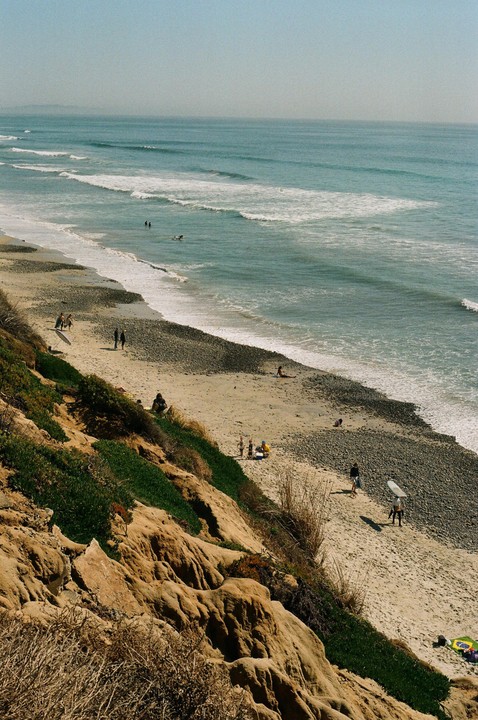 A sandy beach with a rocky shore is visible from a high vantage point, with people on the sand and in the water.