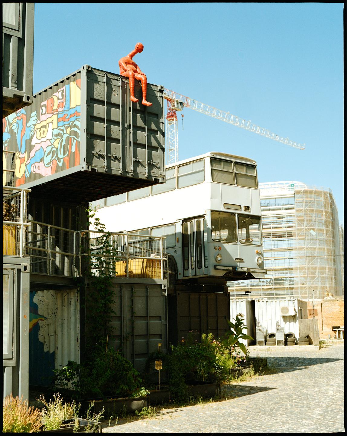 A red sculpture of a man sits atop a stack of shipping containers next to a bus.