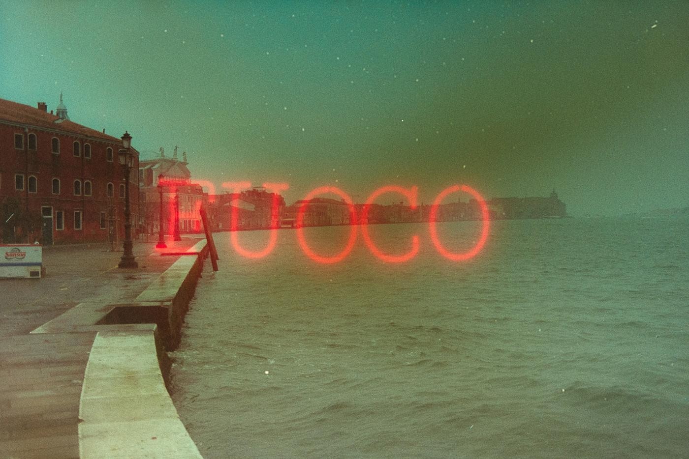A red neon sign spelling "FUOCO" is superimposed over a canal in Venice with buildings along the waterfront.
