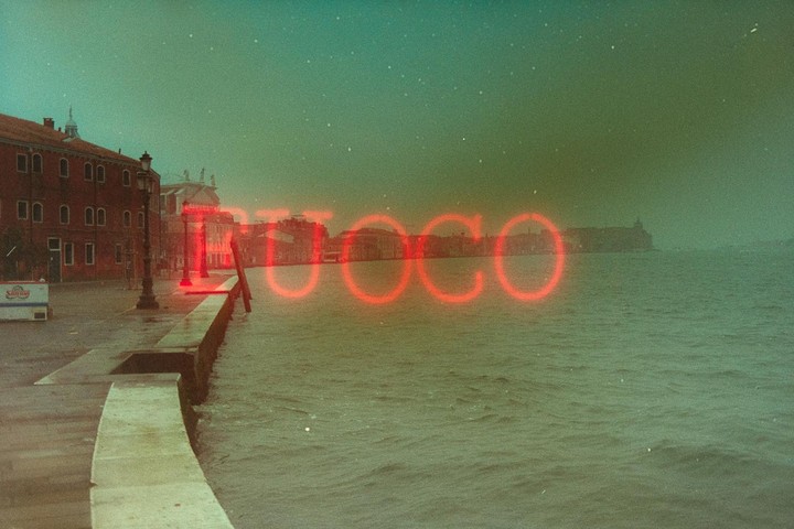 A red neon sign spelling "FUOCO" is superimposed over a canal in Venice with buildings along the waterfront.