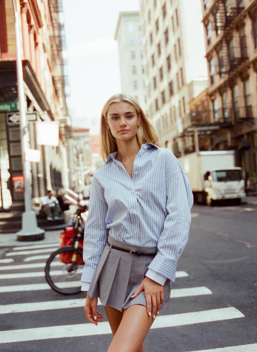 A woman wearing a striped shirt and pleated skirt stands in the middle of a city street.