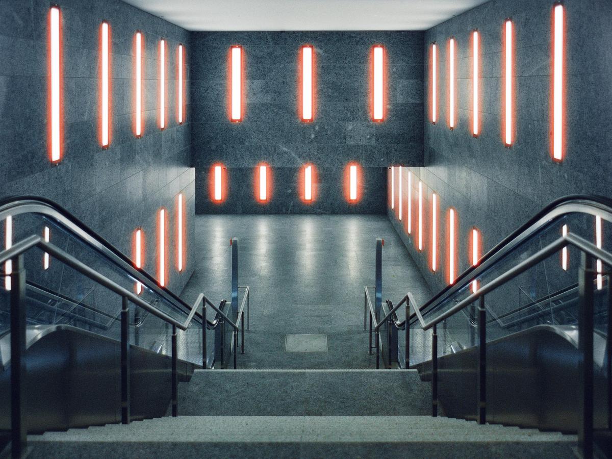 An empty subway station features two escalators descending into a brightly lit underground area with red neon lights on the walls.