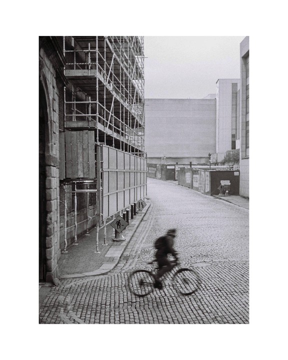 A cyclist rides down a cobblestone street next to a building covered in scaffolding.