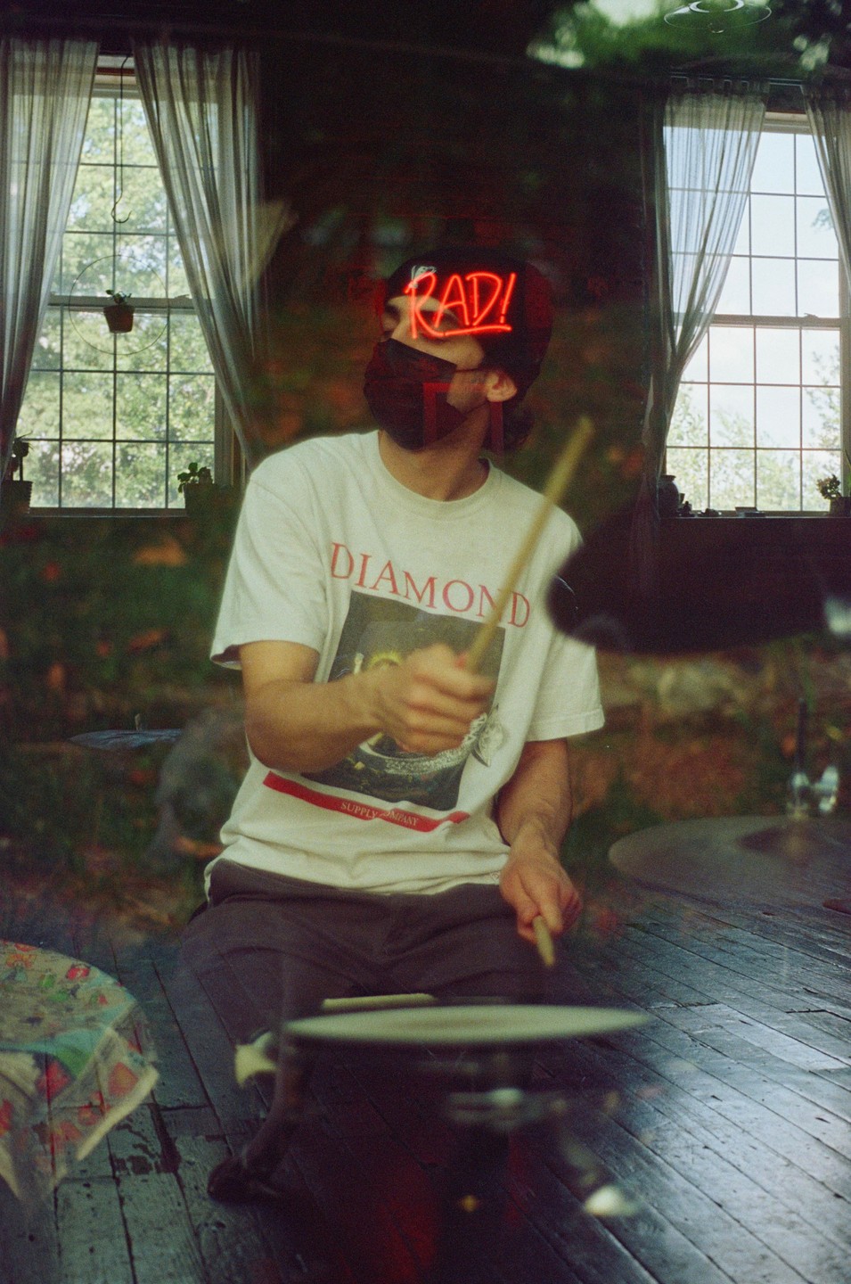 A man wearing a mask and a hat with a neon "RAD!" sign is playing drums in a room with two windows.