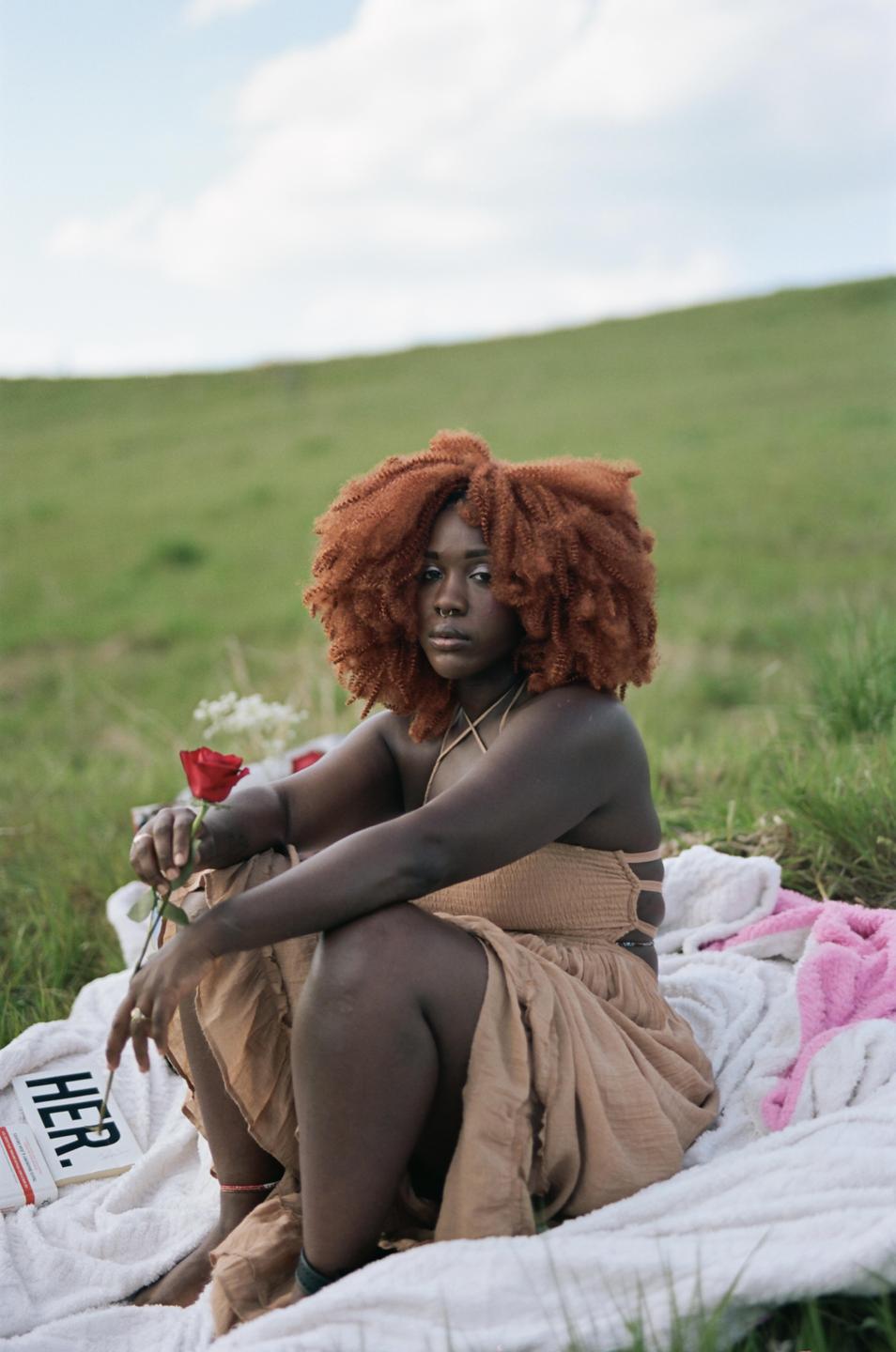 A woman with red hair sits on a white blanket in a grassy field, holding a red rose.