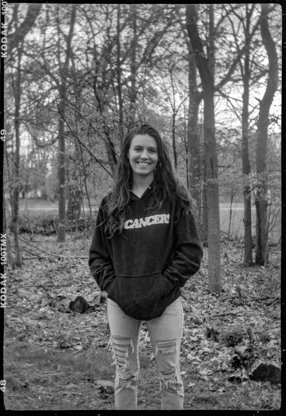 A woman wearing a hoodie and ripped jeans stands in a forest.