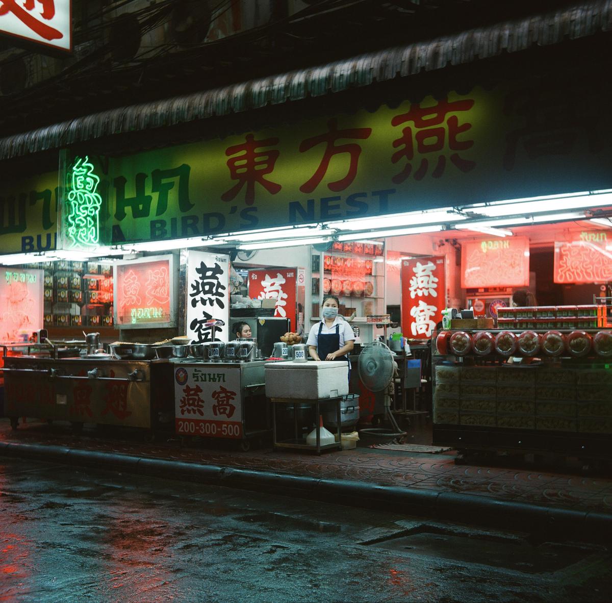 A woman wearing a face mask stands behind a counter at a storefront with neon signs at night.