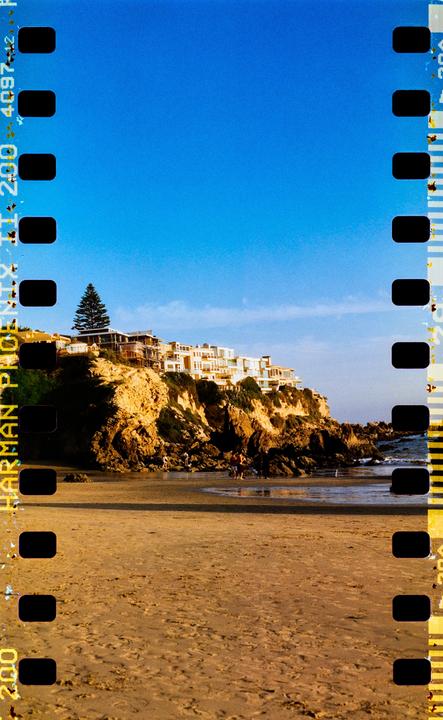 A sandy beach with people walking near the water is overlooked by houses built on a rocky cliff.