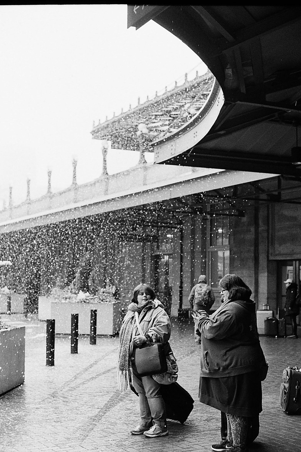 Two women stand outside a building entrance as snow falls heavily around them.