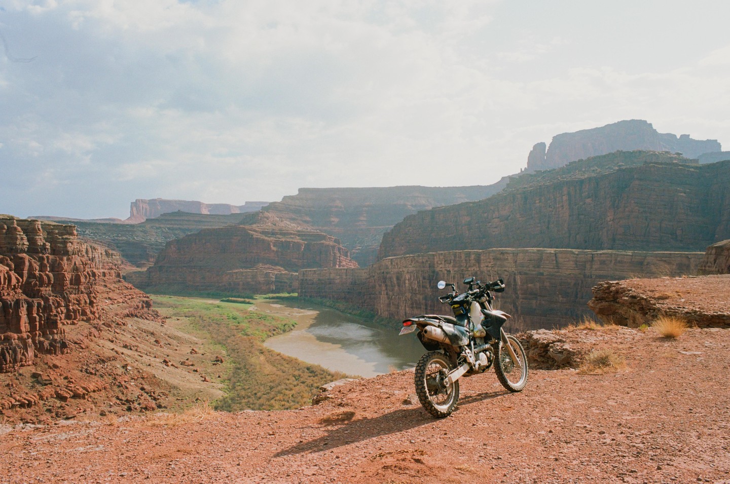 A motorcycle is parked on a dirt road overlooking a river winding through a canyon.