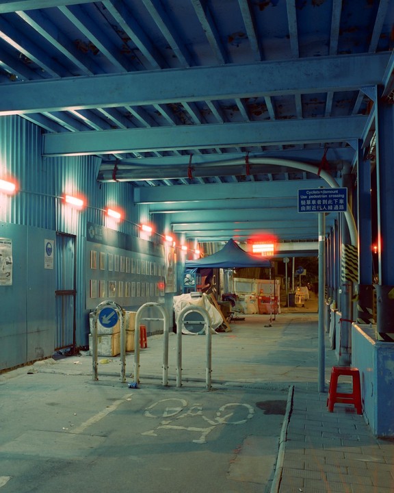 A blue metal structure with red lights extends over a street with a bicycle parking area and a blue tent.