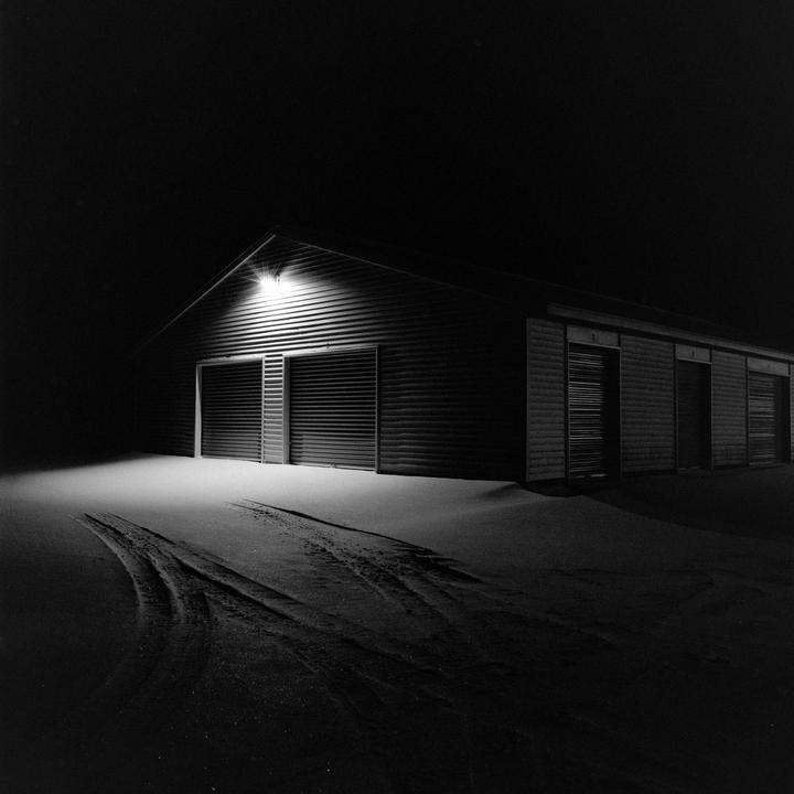 A building with multiple garage doors is illuminated by a single light at night, with snow covering the ground and tire tracks visible.