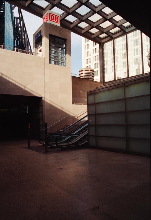 A modern building with a glass roof and escalators is shown with strong shadows.