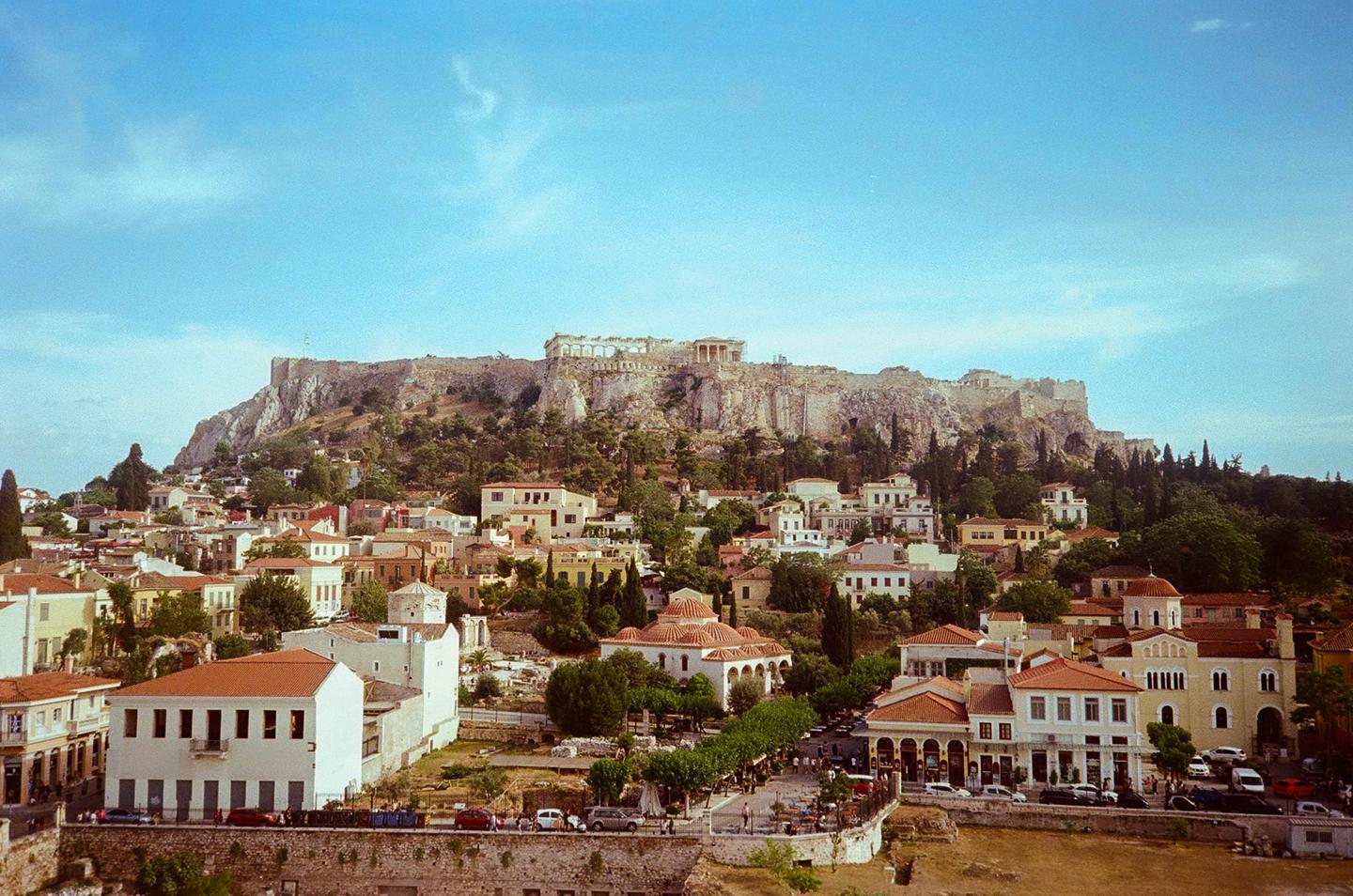 A wide view shows the Acropolis of Athens towering over the Plaka neighborhood, with many buildings and trees in the foreground.