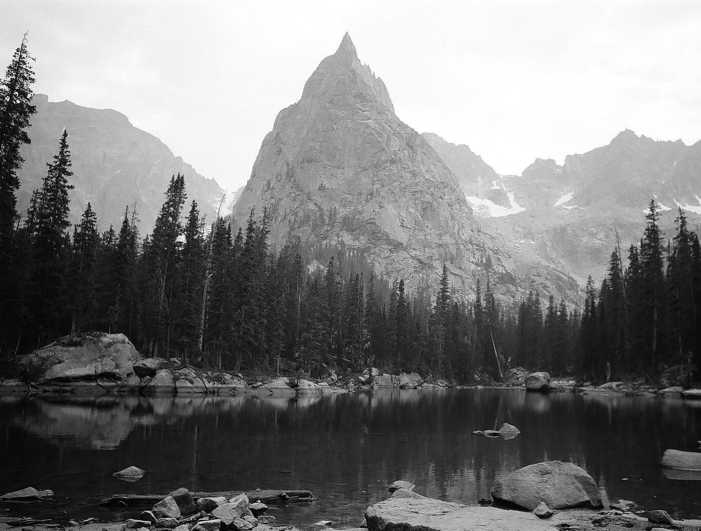 A lake reflects a forest and a large mountain under a cloudy sky.