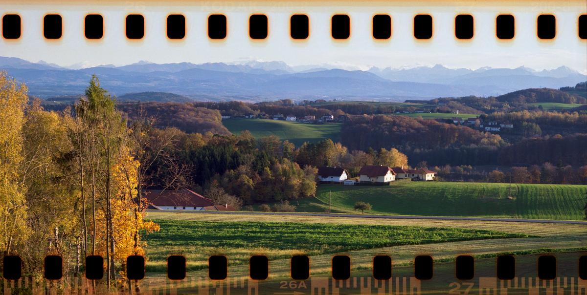 A rural landscape with houses and fields in the foreground, autumn trees in the middle ground, and mountains in the distance.