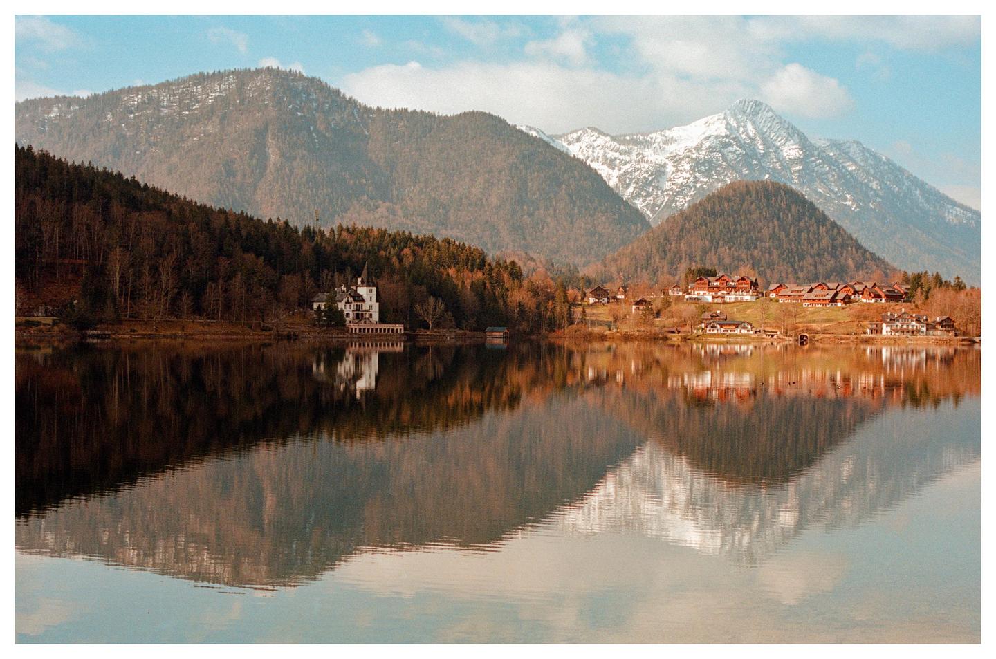 A white castle and a village are reflected in a lake surrounded by mountains.