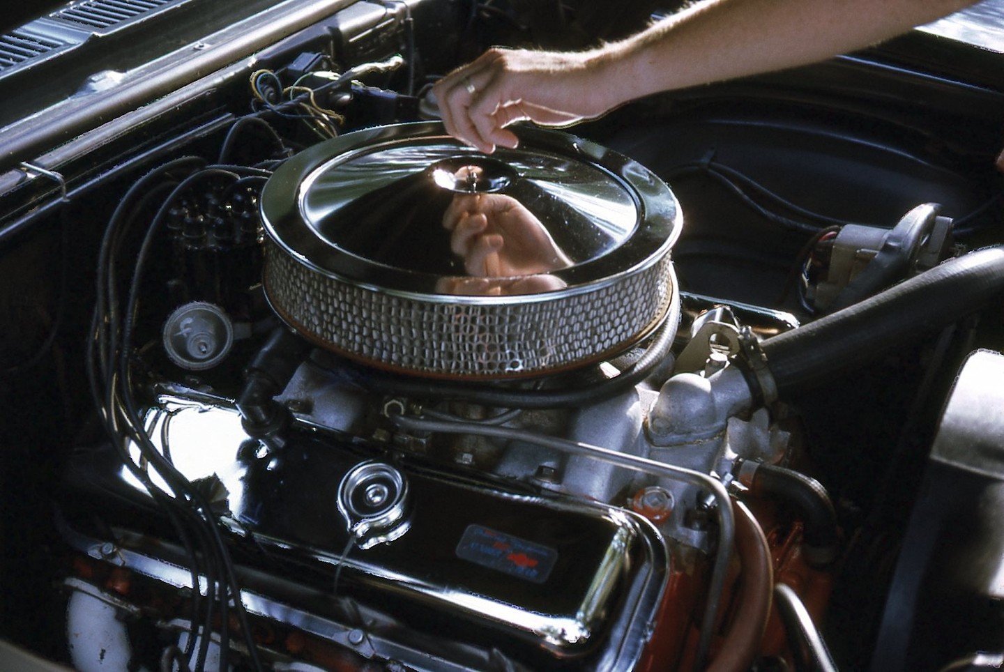 A person's hand is visible above a car engine with a chrome air filter.