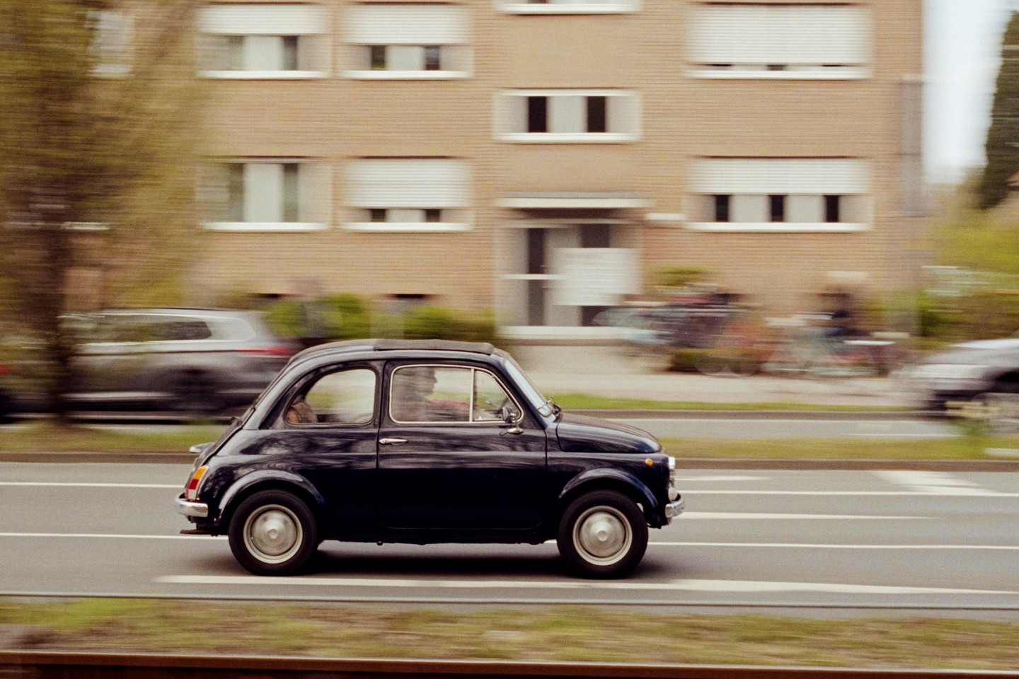 A dark blue Fiat 500 drives past a building with many windows and white blinds.