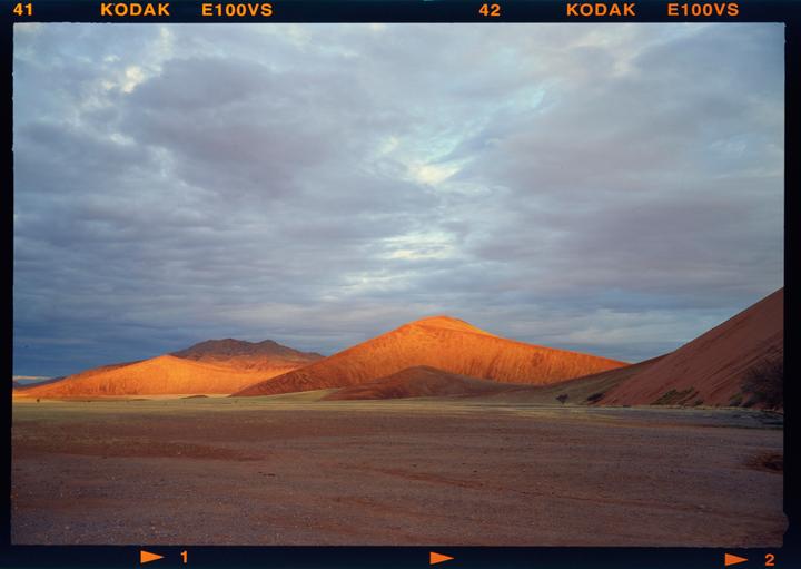 A vast desert landscape features large orange sand dunes under a cloudy sky.