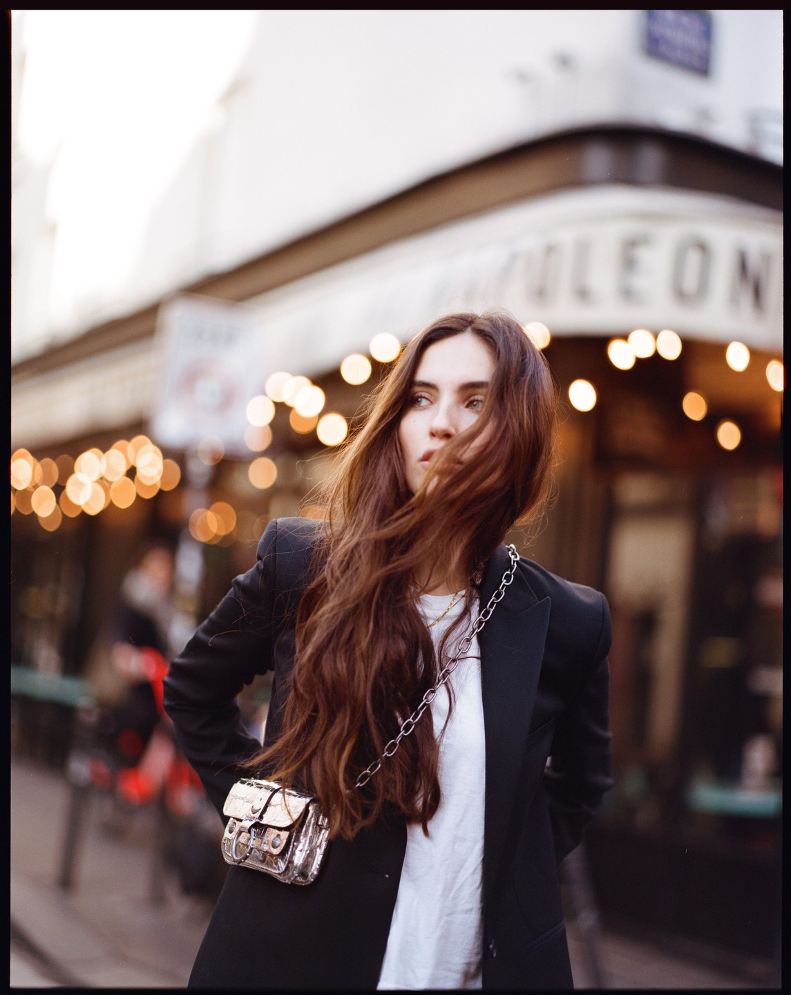 A woman with long brown hair and a black blazer stands on a street with a building and lights in the background.