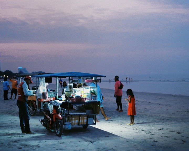 A child in a red dress stands on a beach next to a food cart at dusk.