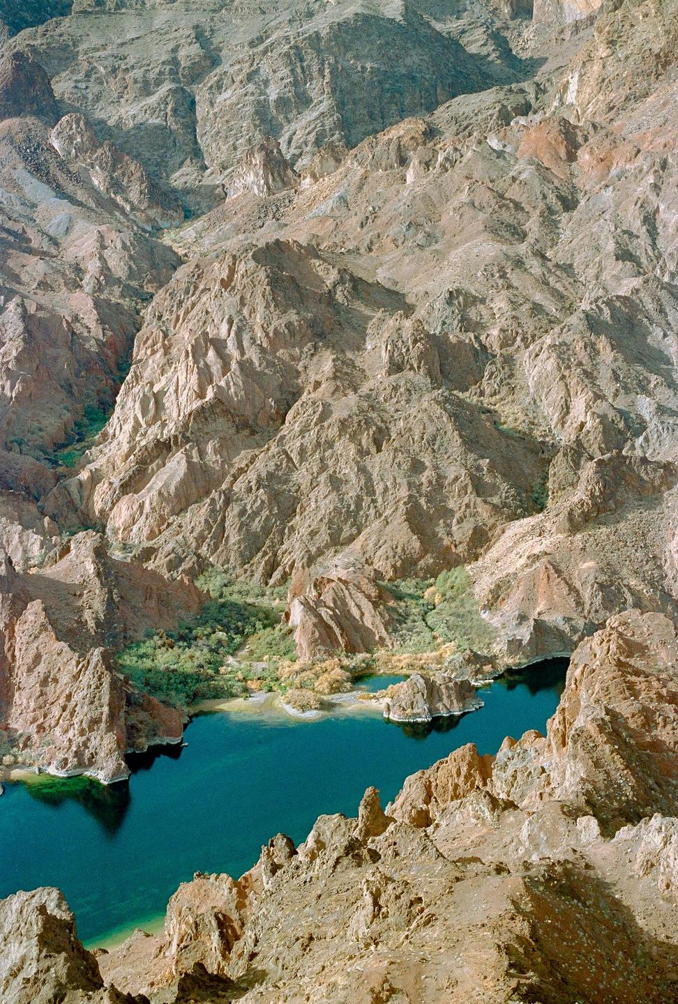 A deep blue lake is nestled among rocky desert mountains with green foliage along its banks.