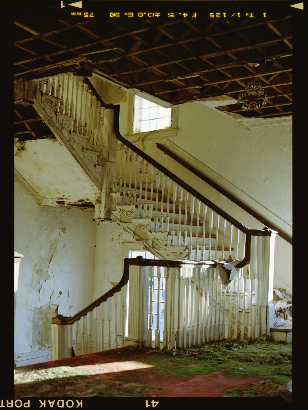 A grand staircase with peeling paint and a damaged ceiling is illuminated by natural light from a window.
