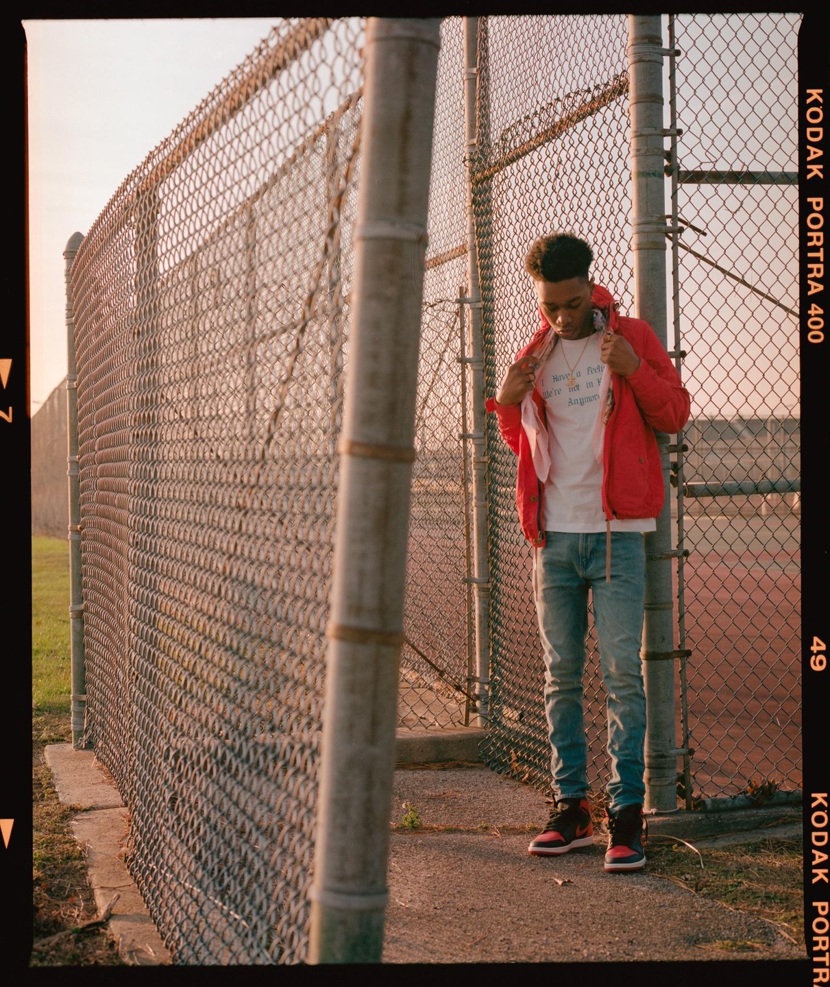 A young man stands next to a chain link fence, adjusting his red jacket.