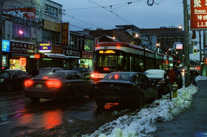A streetcar drives down a city street at night, with cars parked on the side of the road and snow on the ground.