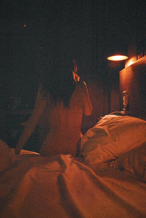 A woman sits on a bed in a dimly lit room with a lamp providing warm light.