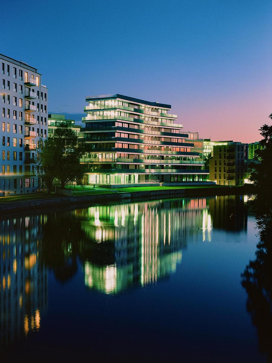 Modern buildings with illuminated windows are reflected in the water at dusk.