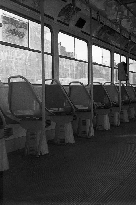 The interior of an empty bus or tram is seen from the back, looking forward, with rows of seats and windows showing buildings outside.
