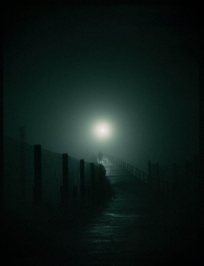 Two people walk on a foggy path at night, illuminated by a bright light in the distance.