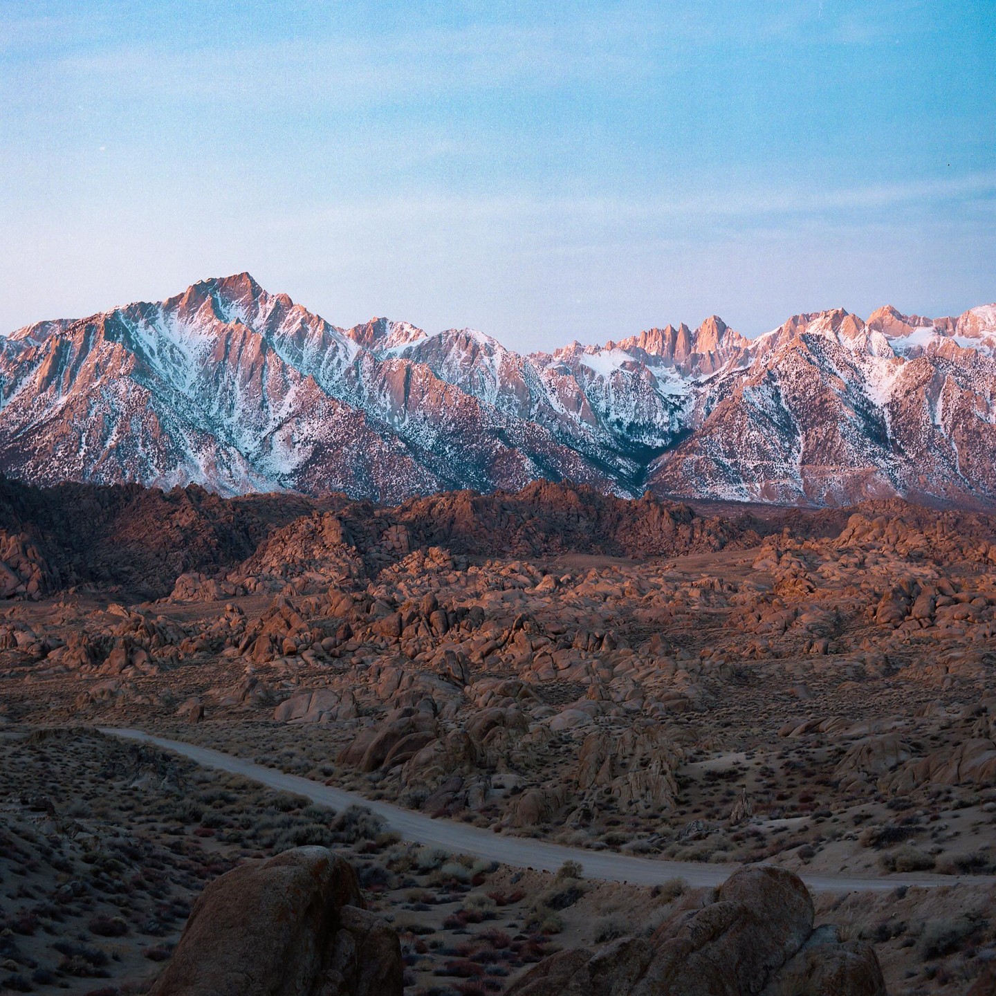 A dirt road winds through a rocky desert landscape with snow-capped mountains in the background under a clear sky.