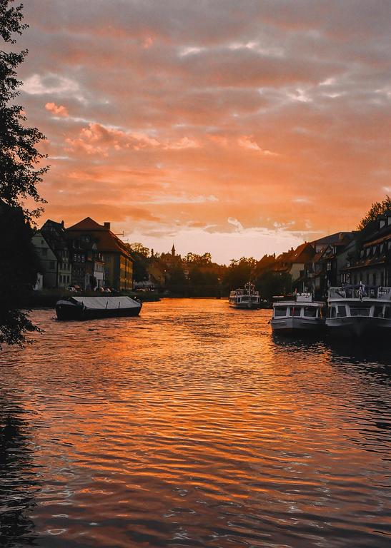 A river with boats and buildings on its banks is illuminated by the orange light of sunset.