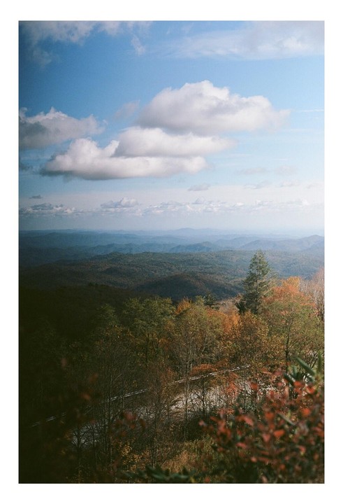 A vast landscape of rolling mountains and hills covered in trees with autumn foliage is visible under a blue sky with white clouds.