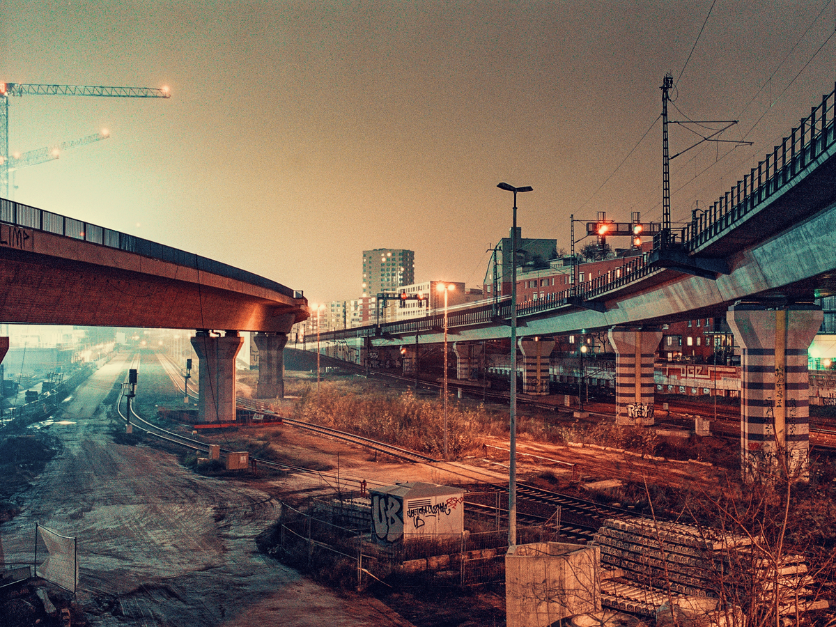 Two elevated train tracks and a highway overpass converge over a network of ground-level tracks and dirt roads at night.