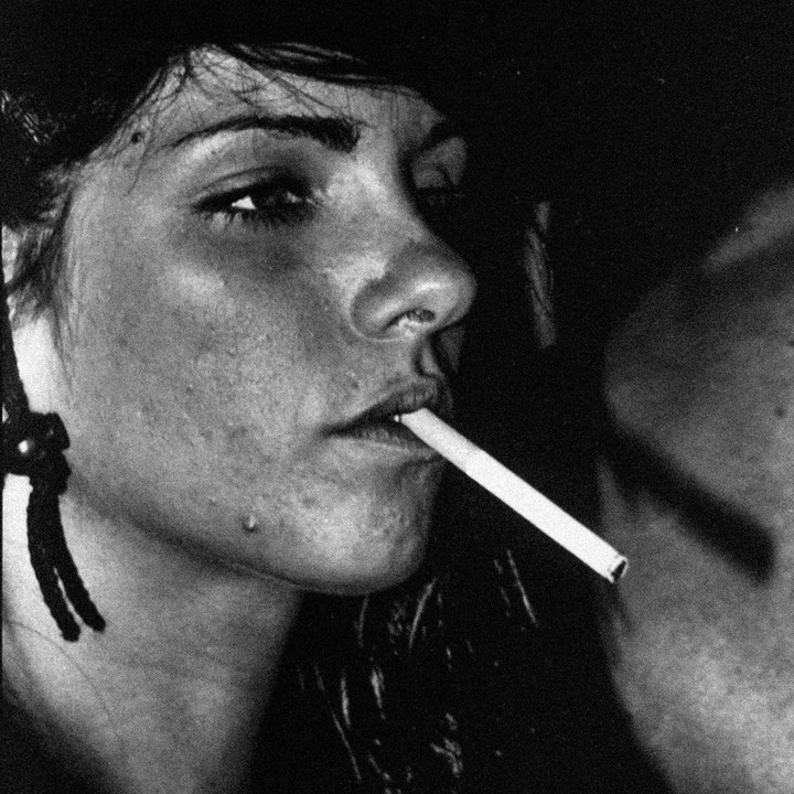 A woman with a cigarette in her mouth is shown in a close-up portrait.
