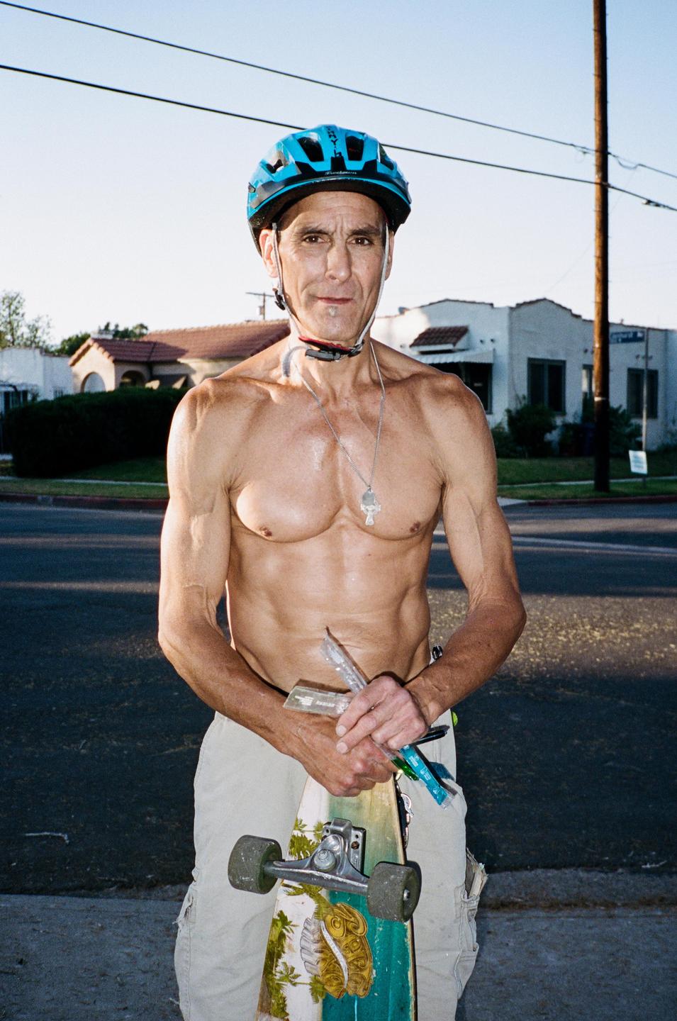 A shirtless man wearing a blue helmet and holding a skateboard stands on a street.