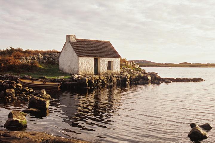 A small white cottage with a brown roof sits on a rocky shore next to a body of water, with a wooden boat moored nearby.